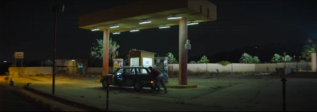 A car and two figures beneath the fluorescent canopy of a quiet petrol station at night.