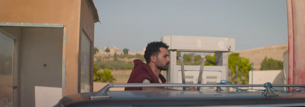 A man in profile beside a fuel pump, with a car roof in the foreground.